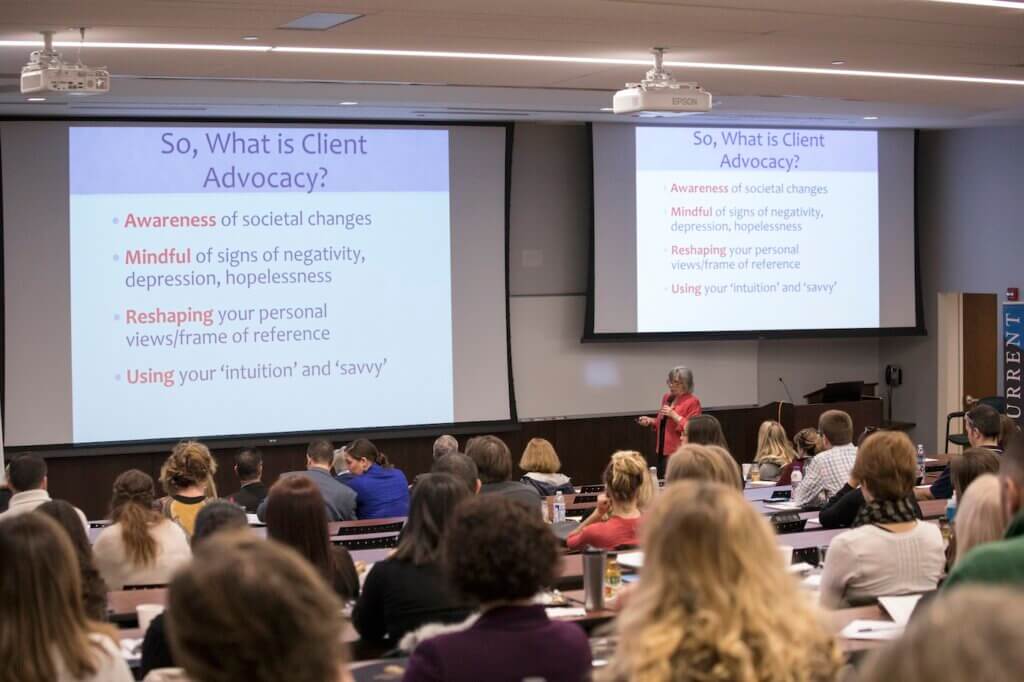 Woman speaking in a packed lecture hall. On two projector screens, the following text appears: "So, what is client advocacy? Awareness of societal changes. Mindful of signs of negativity, depression, hopelessness. Reshaping your personal views/frames of reference. Using your 'intuition' and 'savvy'