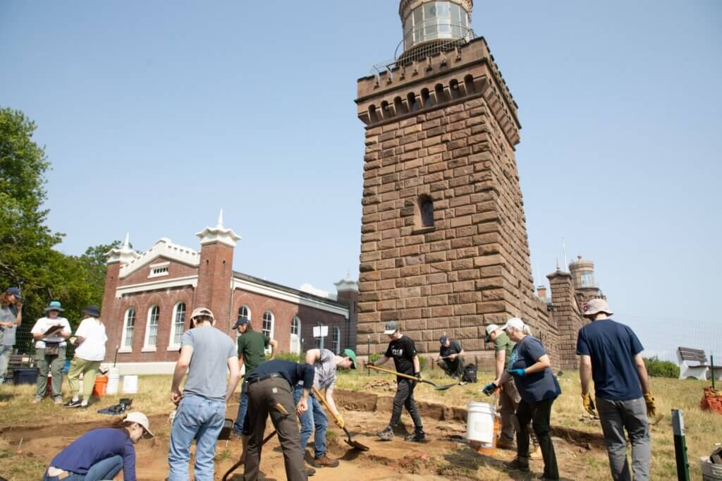 Excavation team digging in front of a light house