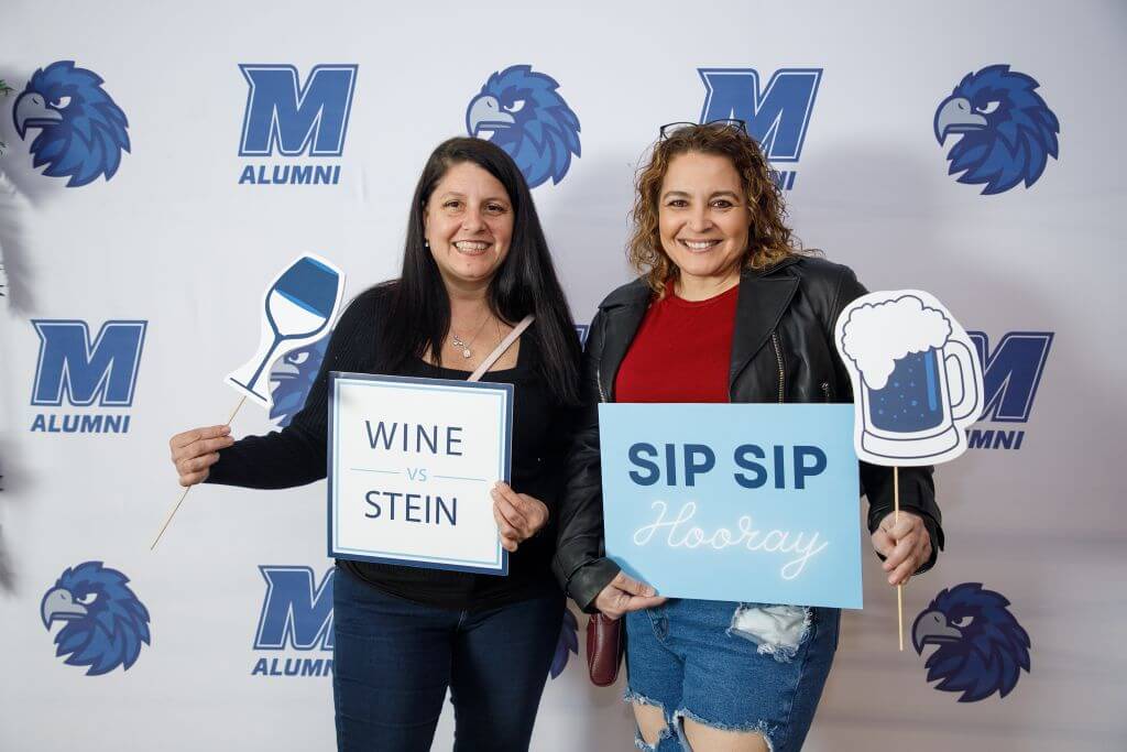 Two Wine vs. Stein participants posing in front of a Monmouth branded banner, holding objects representing a beer mug and a wine glass.