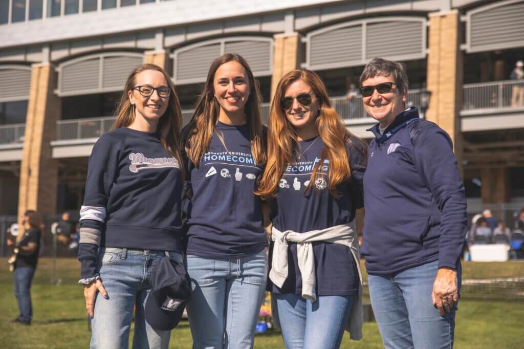 Four women wearing monmouth branded clothing stand in front of the football stadium on a sunny fall day.