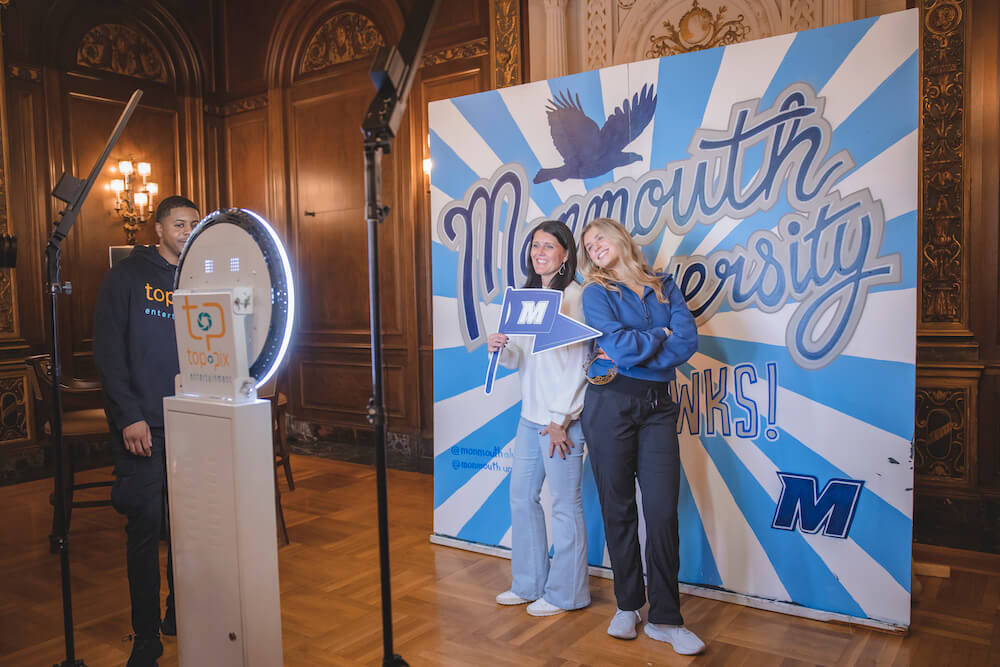 Two woman posing for a shot in front of a mural that says Monmouth University Go Hawks