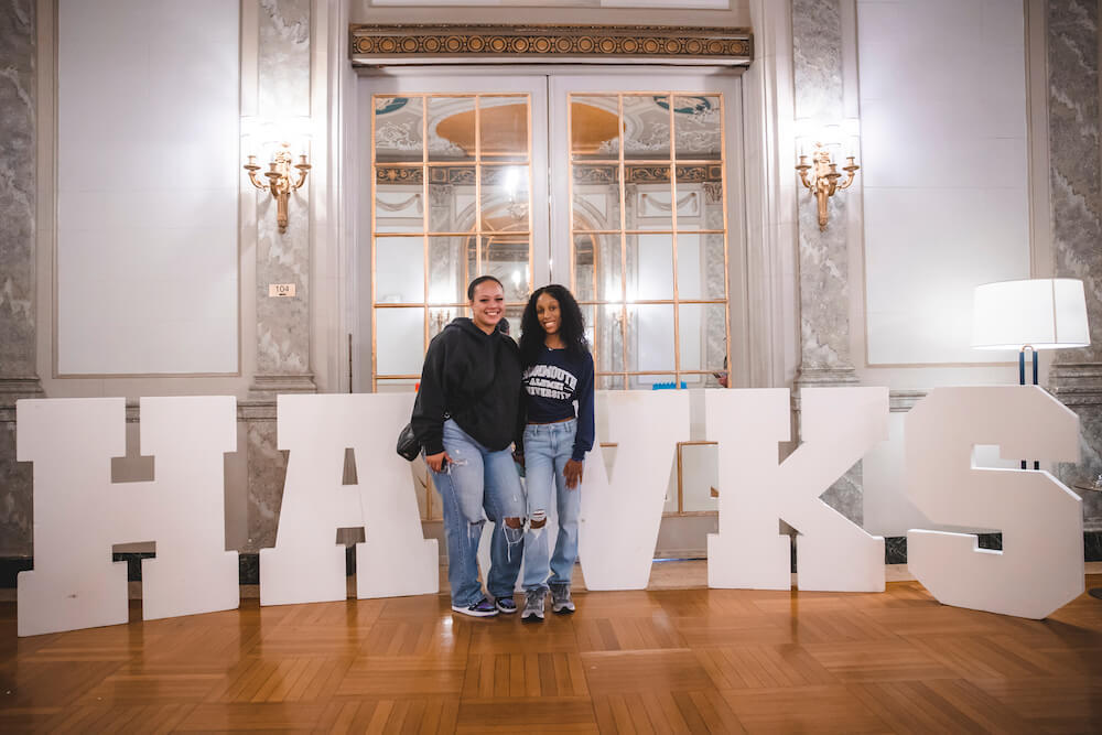Two homecoming attendees posing for a photo in the Great Hall in front of letters spelling out HAWKS
