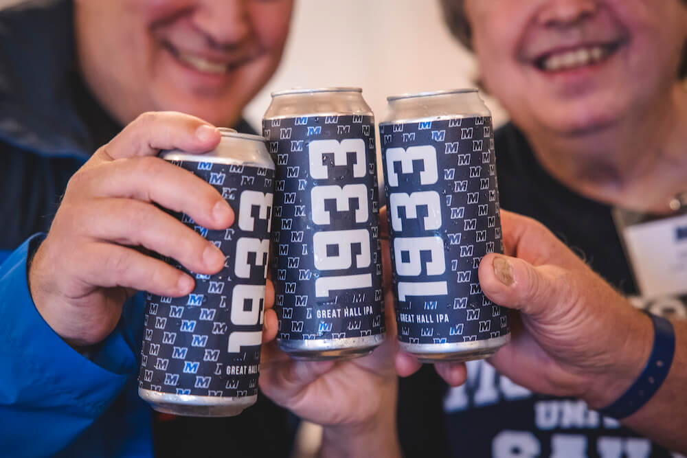 Alumni clinking beer cans together. The can is of Great Hall IPA, labelled 1933.