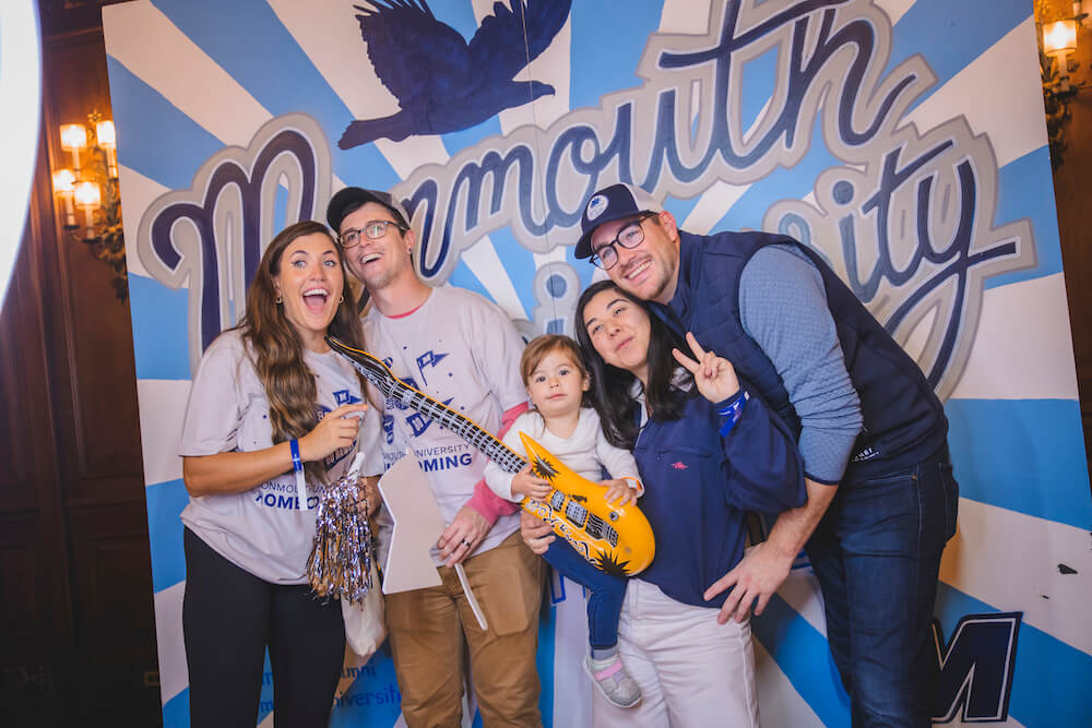 Family posing for a shot in front of a mural for Monmouth University, child holding an inflatable guitar