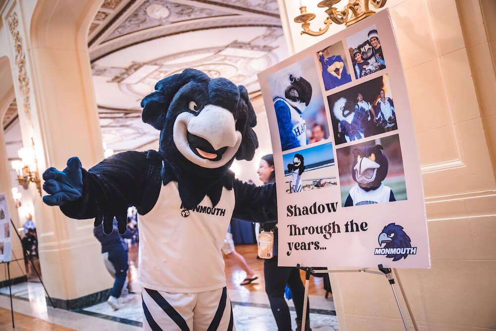 Shadow the Hawk posing for a photo next two a sign, which shows "Shadow Through the Years", his various costumed iterations