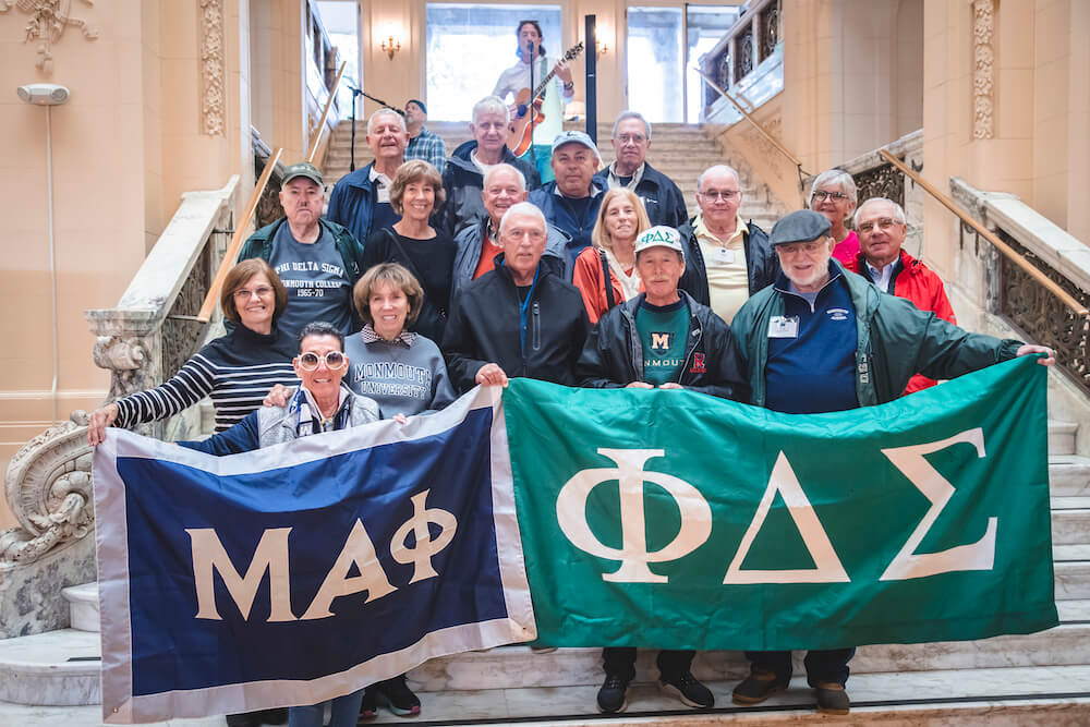 A fraternity and sorority posing for a photo on the indoor steps of the Great Hall, each waving banners showing their greek letters