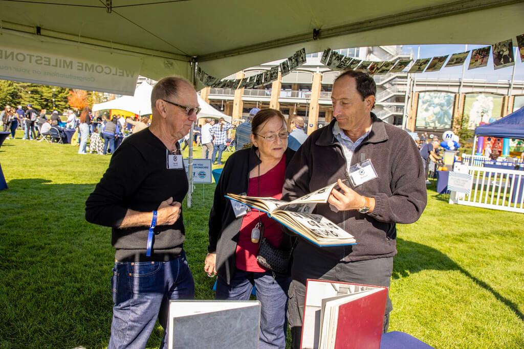 2 men and 1 woman looking at a class of 1975 yearbook at the Homecoming Tailgate Milestone tent