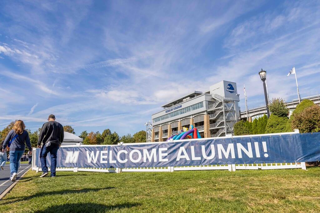 Monmouth "Welcome Alumni!" banner at the Homecoming Tailgate with Kessler Stadium and blue sky in the background.