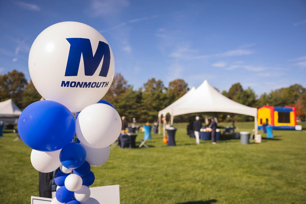 Balloon column with white and blue ballons and the large top balloon with the Monmouth logo. Homecoming tailgate tent and kids bouncehouse in the background.