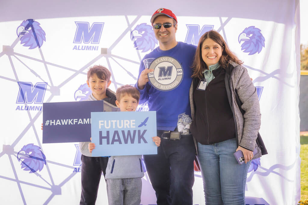 Mom, dad and 2 sons at Milestone Tent. Dad holding up Class of 2000 25th reunion sign and sons holding up "#Hawkfamily" and "Future Hawk" signs
