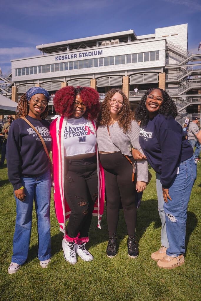 Group of 4 young ladies in Monmouth gear smiling in front of Kessler Stadium
