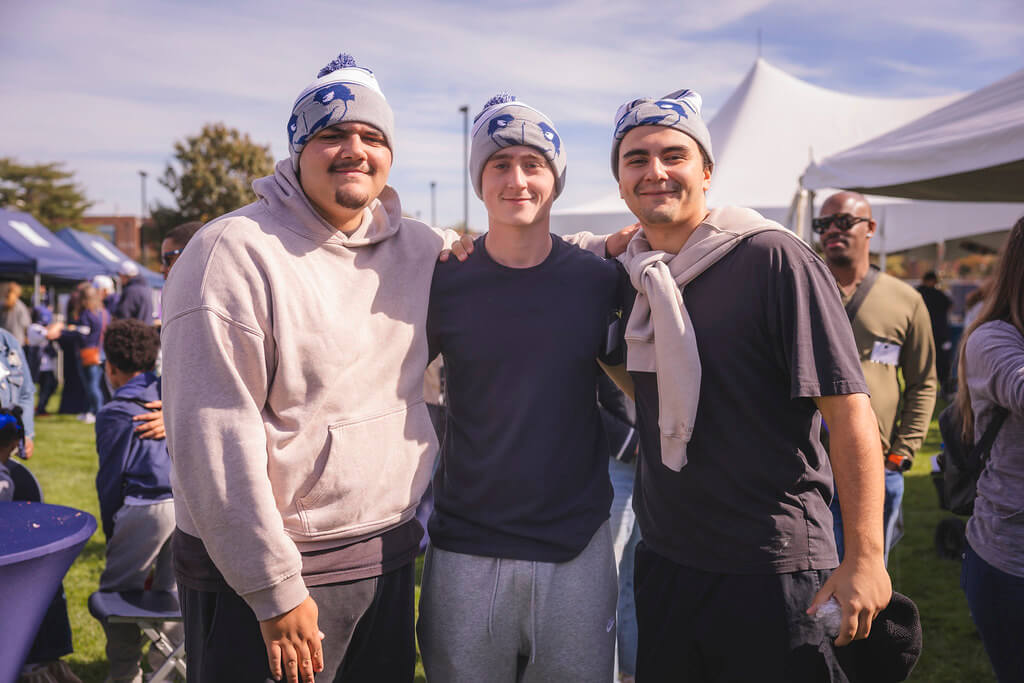 3 young men with their arms around each other wearing the limited edition Homecoming winter hat