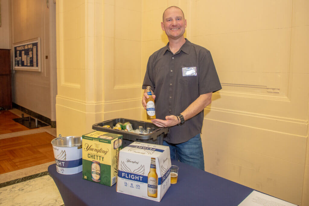 Man holding a bottle of beer and presenting it to the viewer. He stands behind a table with a cooler, an ice bucket, and two cases of beer.