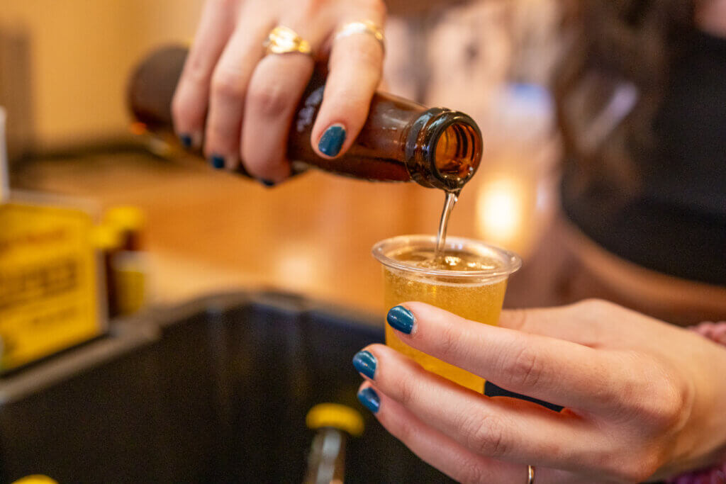 A close-up of a volunteer pouring beer from a bottle into a small plastic tasting cup.
