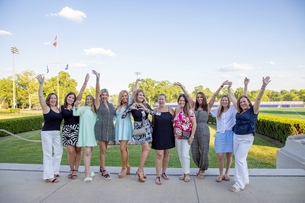 11 women on the lawn of the Great Hall, hands raised in expression of joy. Woman in the center makes a heart shape with her hands.
