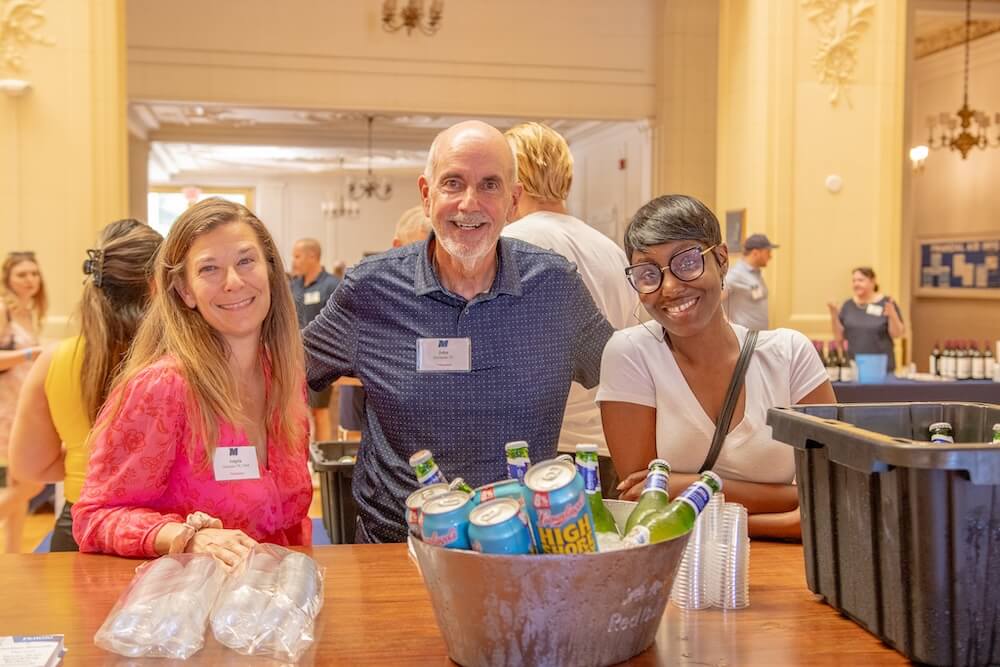 Three wine and stein patrons smiling behind a table with cups and drinks