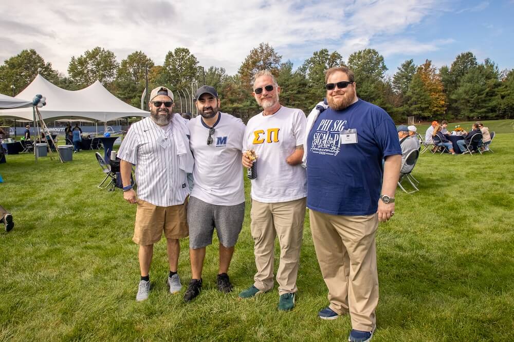Four alumni in sunglasses and Monmouth and Greek-letter shirts pose together on the lawn at the Homecoming tailgate. Tents and guests seated at tables are behind them.