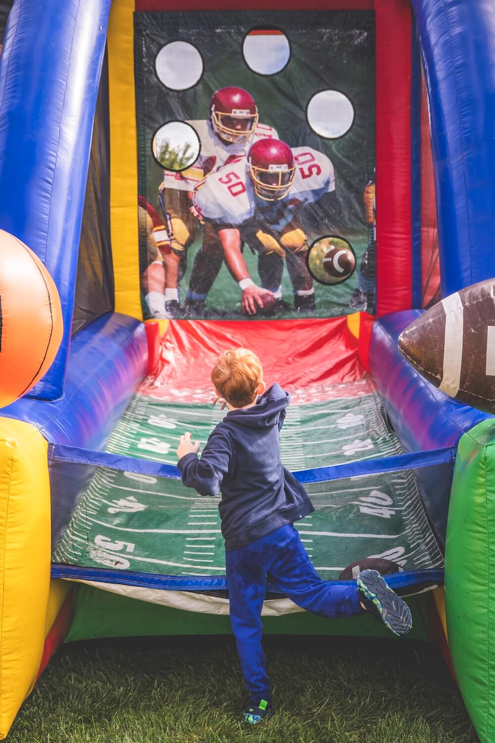 A young child in a navy hoodie throws a football into an inflatable football toss game with target holes at the Homecoming tailgate.