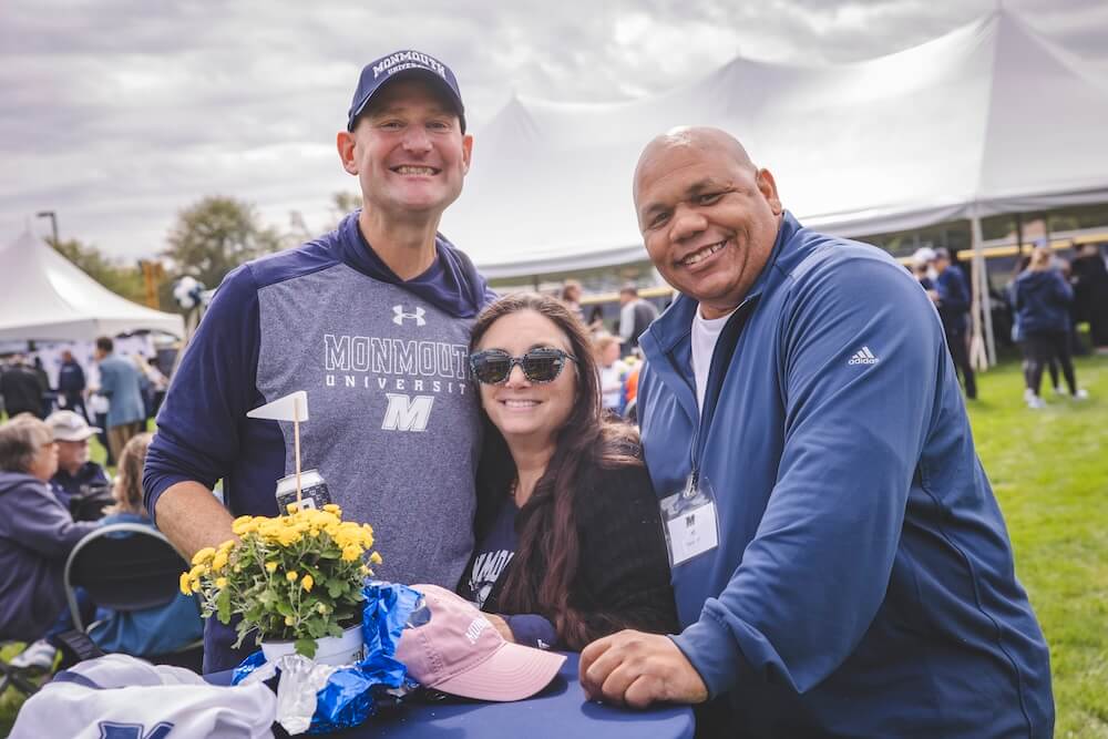Three alumni in Monmouth gear lean on a cocktail table decorated with yellow mums and smile for the camera. Large white tents and the tailgate crowd are behind them.