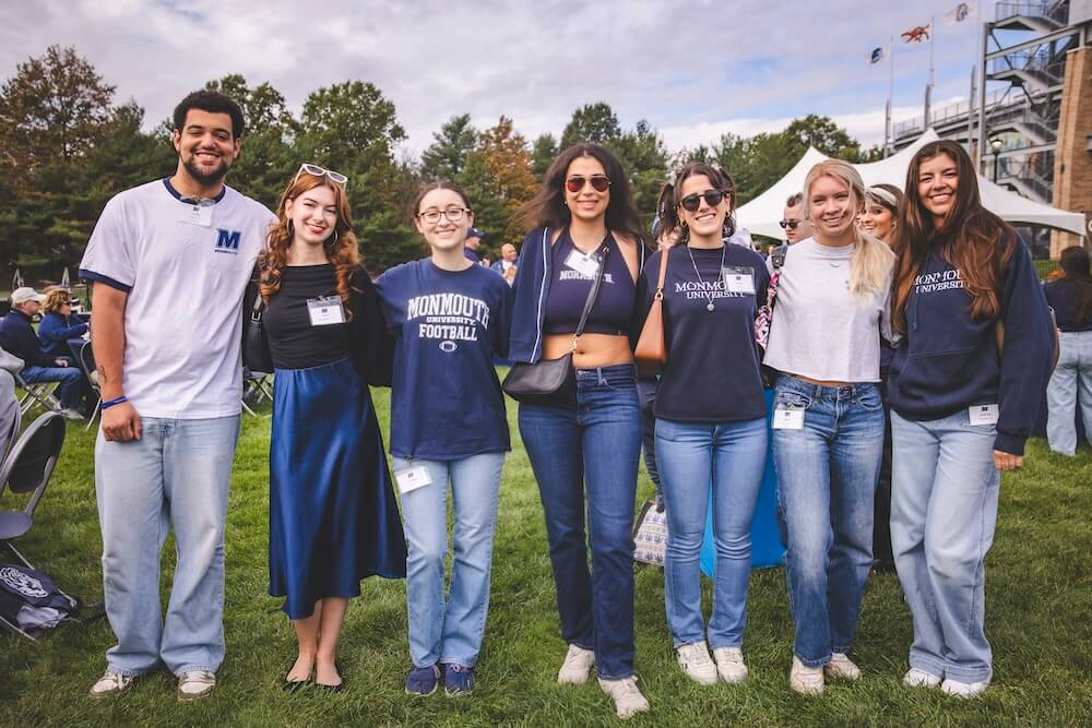 Seven young alumni in Monmouth shirts and jeans stand in a row on the grass, smiling at the Homecoming tailgate, with tents and stadium flags behind them.