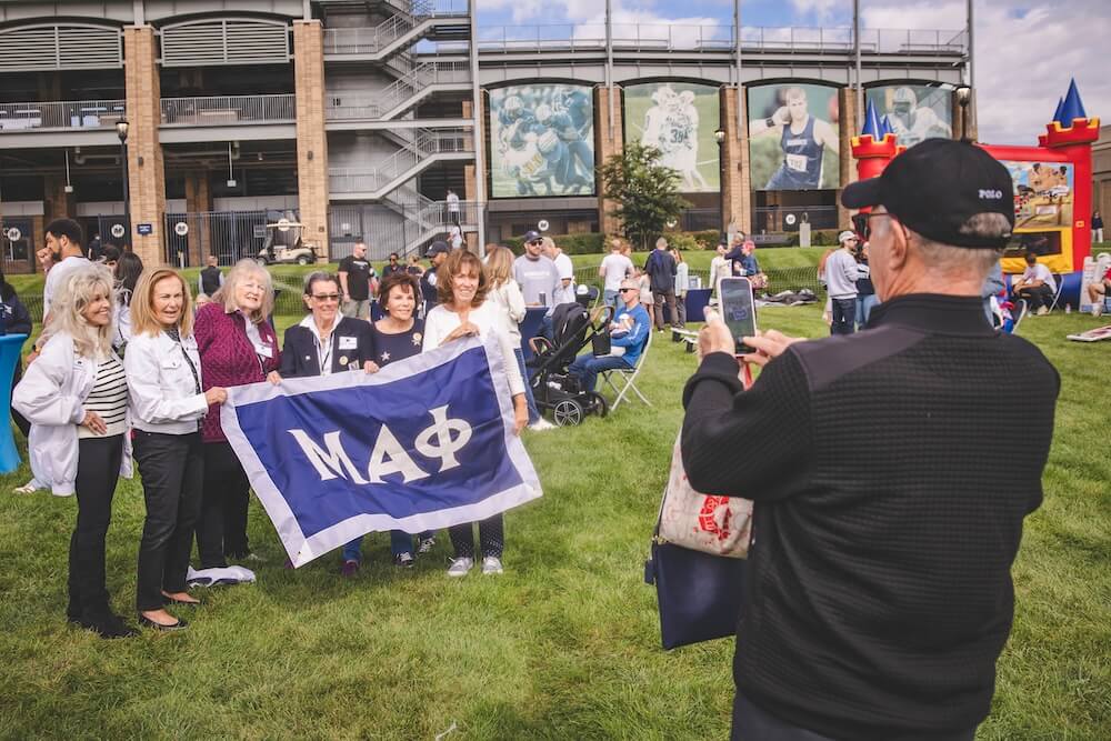 A group of alumni hold up a blue Mu Alpha Phi flag while a guest takes their photo with a phone on the lawn outside Kessler Stadium.