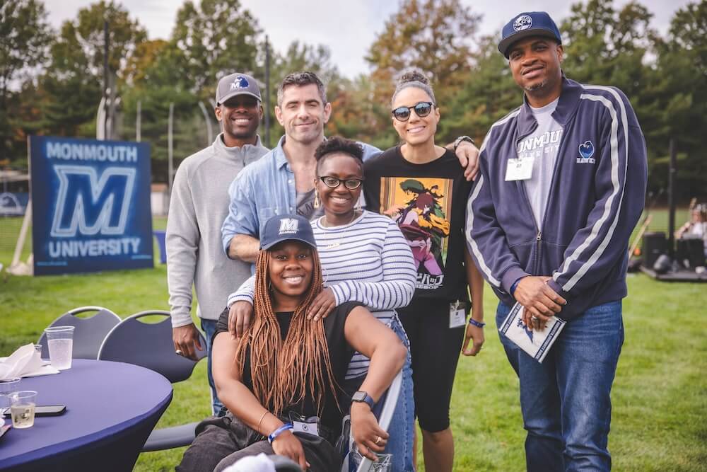 Six friends gather around a table at the Homecoming tailgate and smile for a group photo, with a Monmouth University sign behind them.