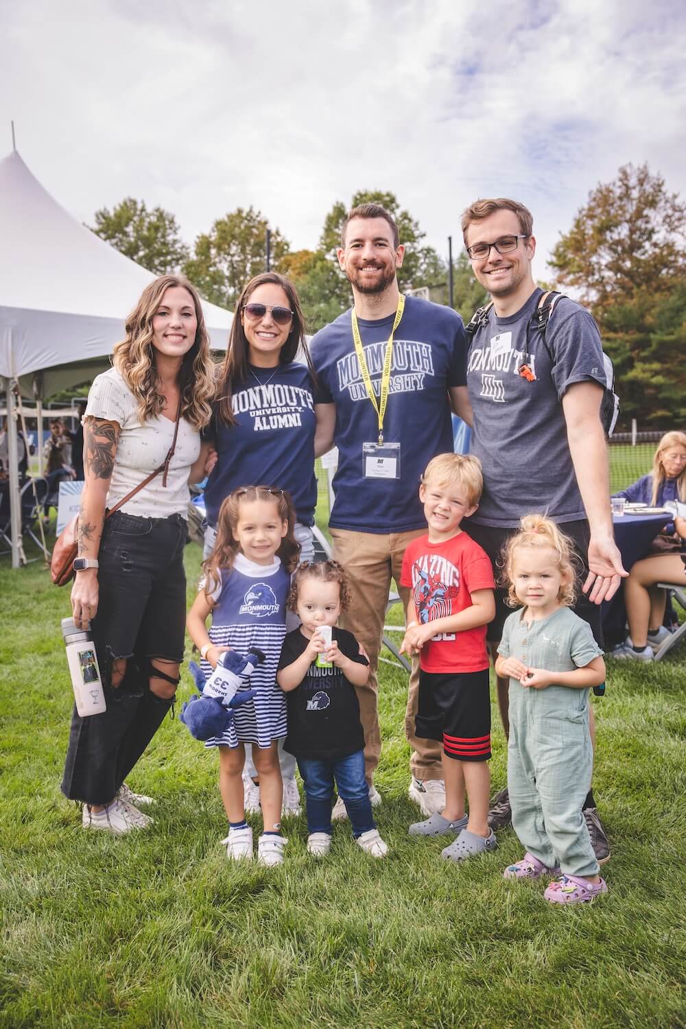 Four alumni in Monmouth shirts pose on the tailgate lawn with four young children, one of whom holds a plush Monmouth Hawks mascot.