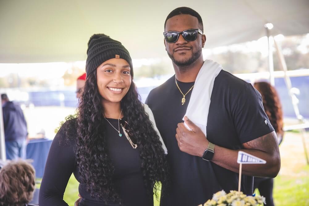 Two guests in black smile for a photo under a tent at the Homecoming tailgate, next to a small “Go Hawks” pennant in a flower centerpiece.