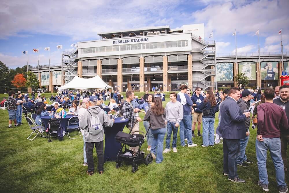 A large crowd of alumni and families mingles at tables and under a white tent on the lawn in front of Kessler Stadium during the Homecoming tailgate.