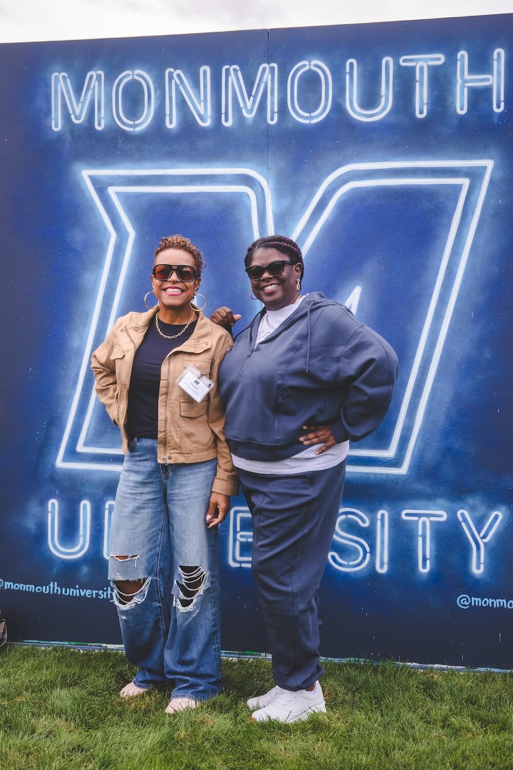 Two alumni in sunglasses pose arm in arm in front of a glowing neon-style Monmouth University mural.