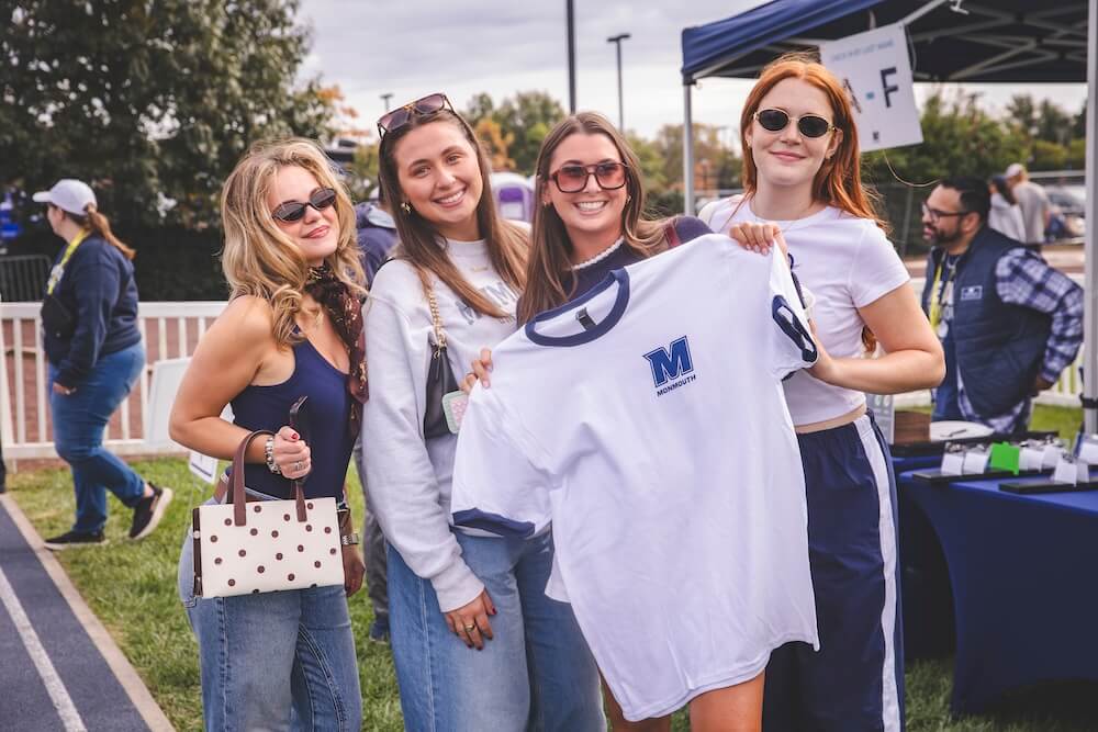 Four young alumni in sunglasses smile at the check-in tent while one holds up a white Monmouth “M” ringer T-shirt.