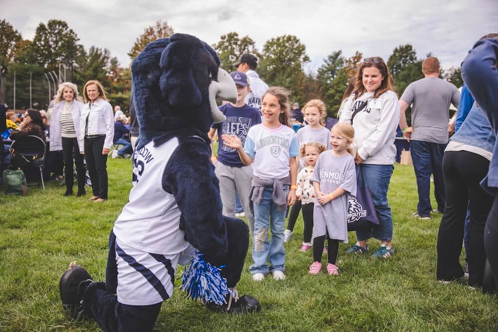 Shadow the Hawk, Monmouth’s mascot, kneels on the grass to greet a group of excited children as families look on at the Homecoming tailgate.