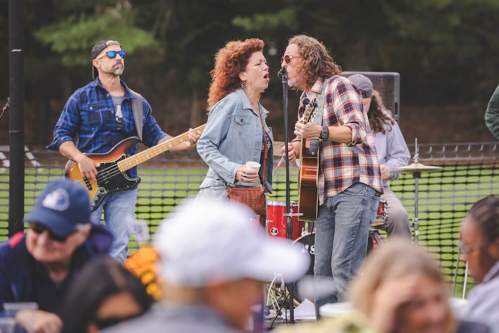 A live band plays at the Homecoming tailgate: two singers share a microphone, one playing acoustic guitar, while a bassist and drummer play behind them.