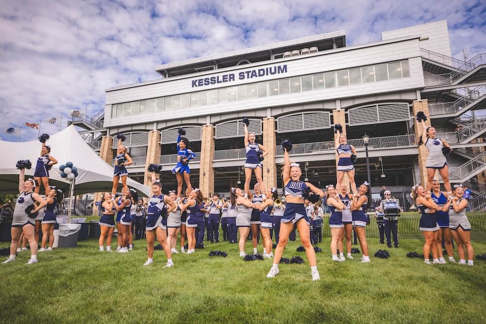 Monmouth cheerleaders perform stunts on the lawn in front of Kessler Stadium, with flyers lifted high and pom-poms raised, as the marching band plays behind them.