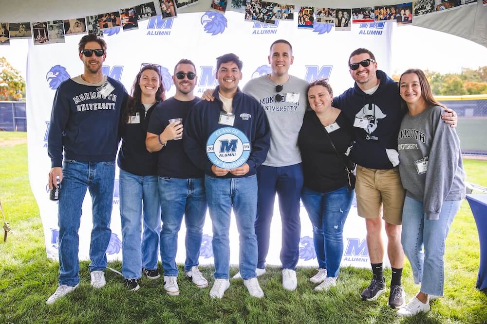 Eight alumni from the Class of 2016 pose in front of a Monmouth Alumni backdrop hung with old photos. The person in the center holds a 10th reunion sign.