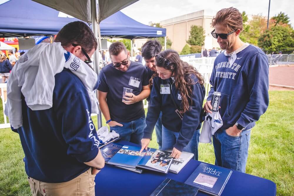 Alumni lean over a table under a tent to look through open Monmouth yearbooks, including a copy of “Tides of Time.”