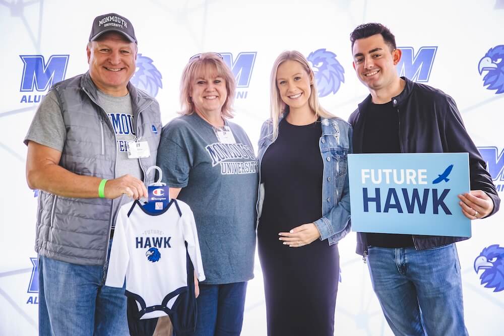 Four guests smile in front of a Monmouth Alumni backdrop, one holding a baby bodysuit and another a sign, both reading “Future Hawk,” while the pregnant guest between them rests a hand on her belly.