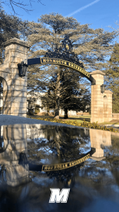 The gated entrance to Monmouth University, with a puddle reflecting it at the lower half of the photo