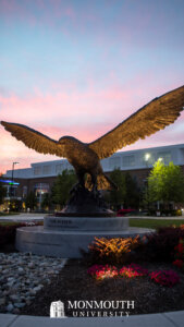 A large hawk statue with its wings stretched out is lit from underneath with a multicolored sunset behind it. 