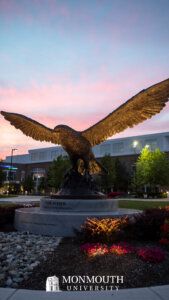 A large hawk statue with its wings stretched out is lit from underneath with a multicolored sunset behind it.