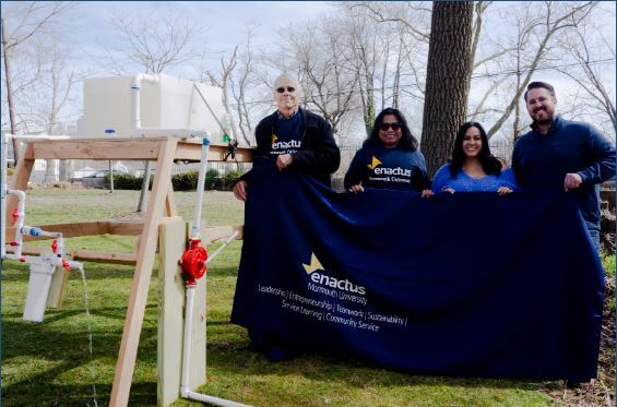 four people stand with a banner for Enactus next to a water filtration system. 