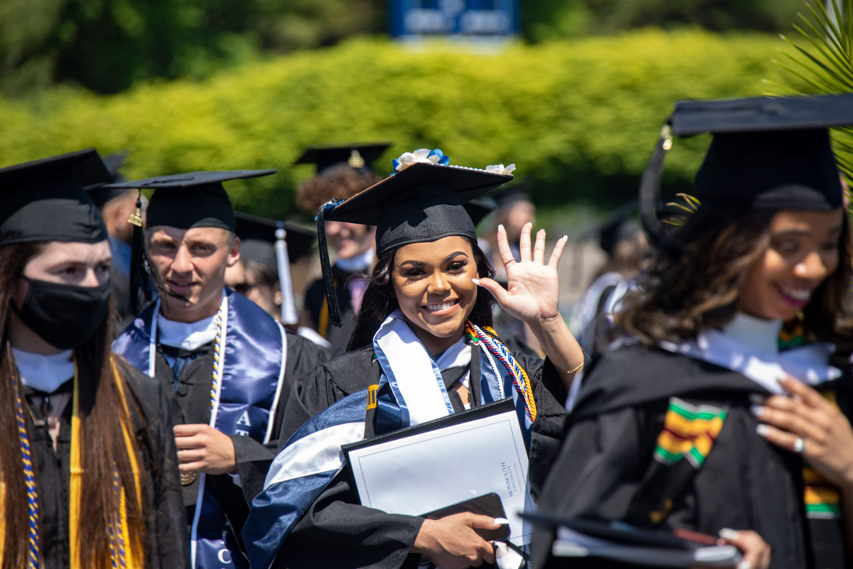 A woman holding a diploma is wearing a commencement gown and cap, she waves at the camera and is surrounded by other smiling graduates