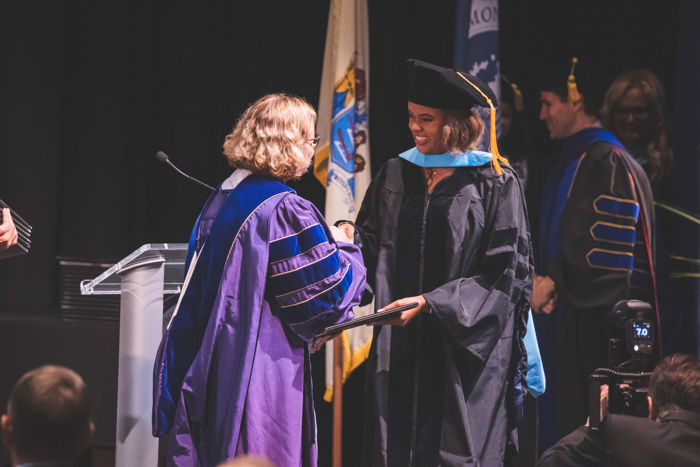 Graduate in cap and gown, smiling while she accepts a degree from a member of the faculty, who is wearing a purple and blue doctoral robe.