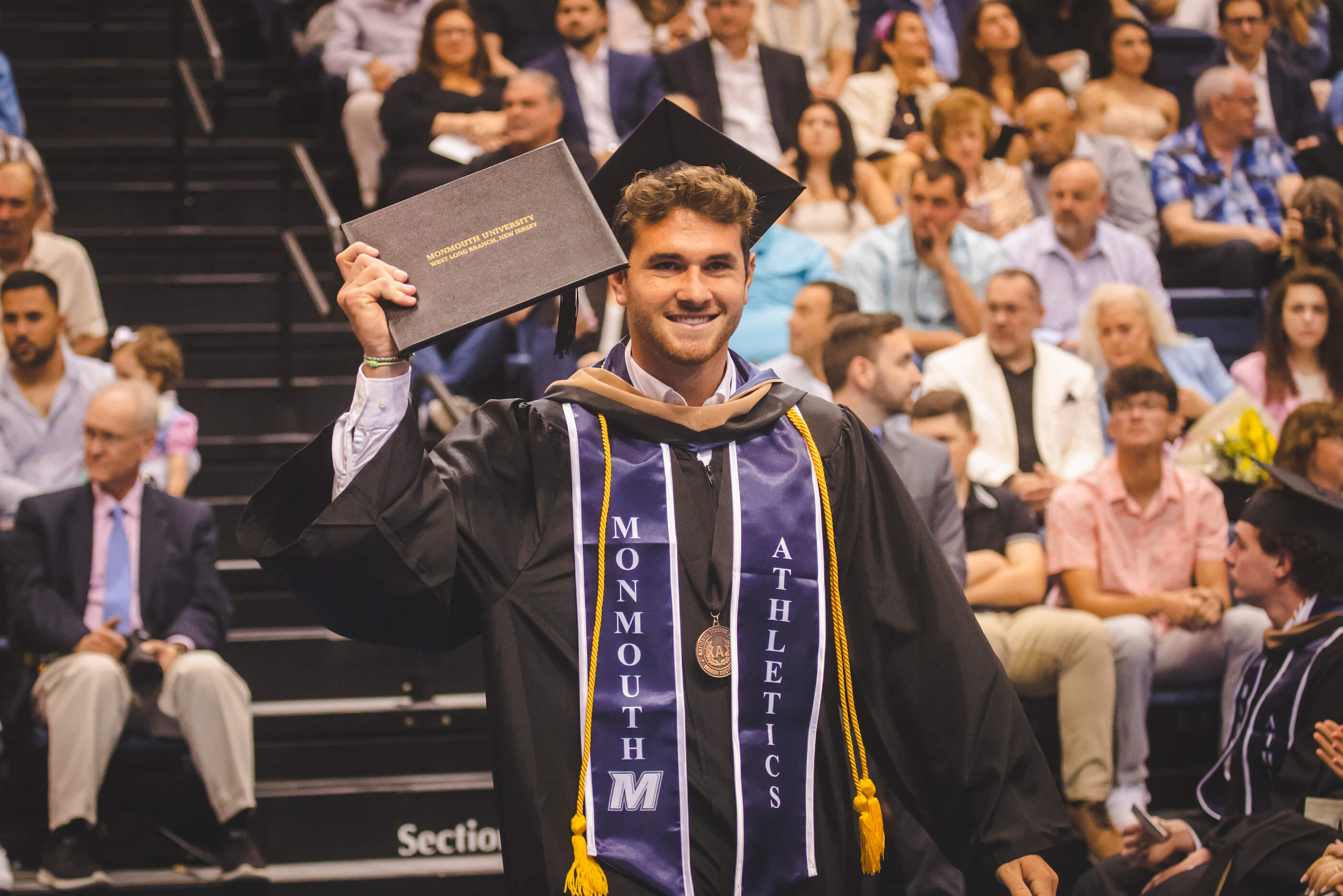 Man in cap and gown holding up the degree he just earned, crowd in the back clapping