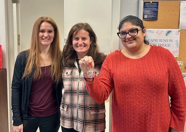 Three women smile for camera, one of whom is holding a holiday ornament