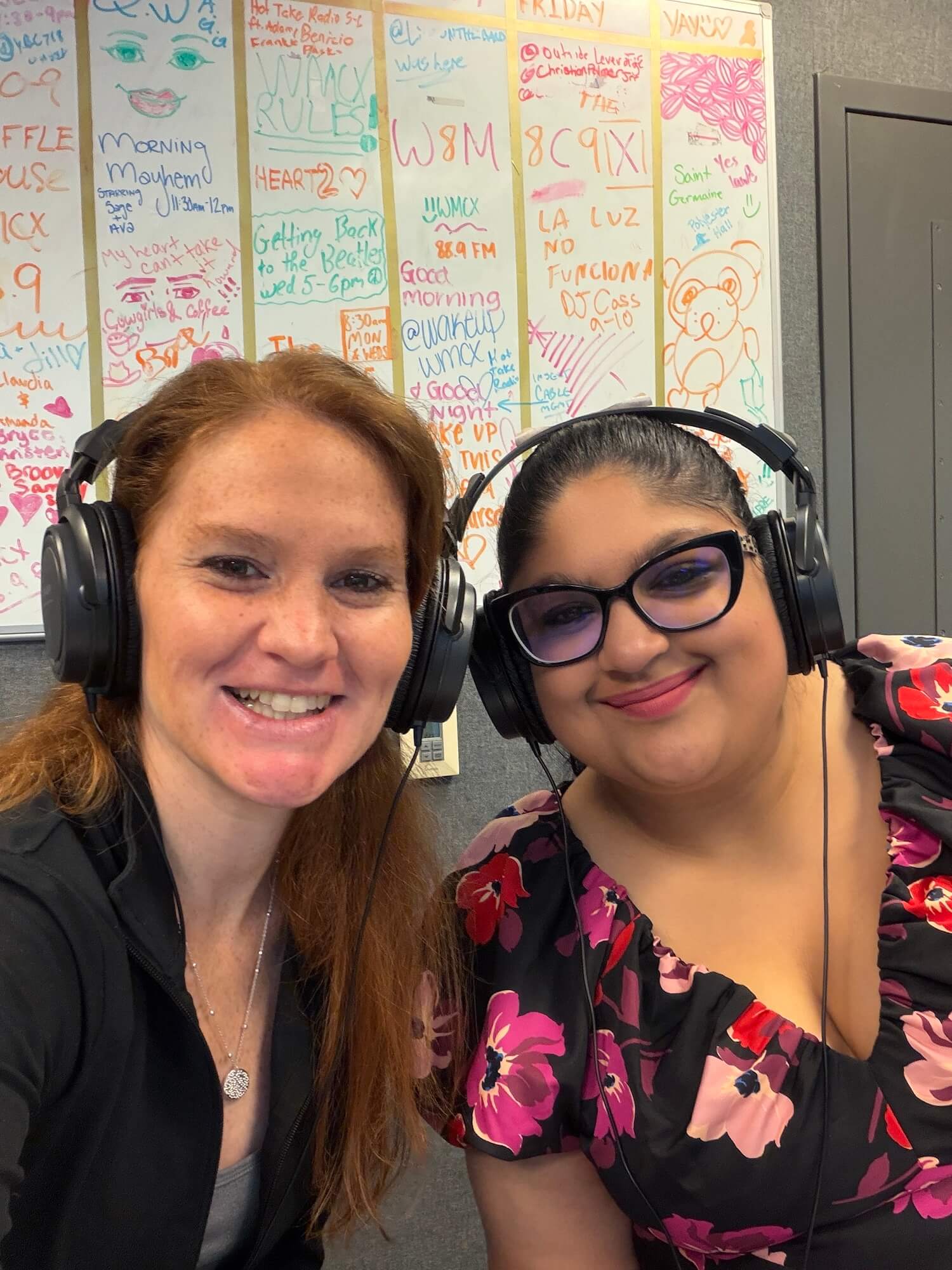 Two women smiling, wearing large headphones. Marker drawings and writings in the back show a schedule for the WMCX radio station.