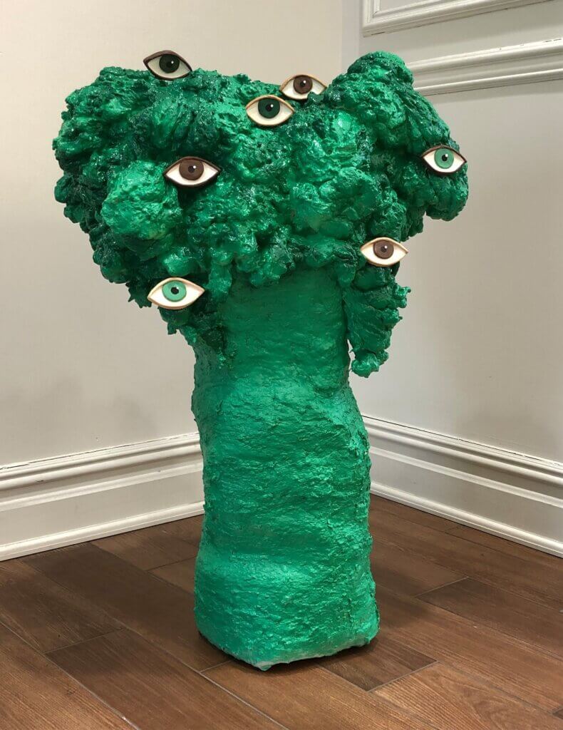 Green sculpture with a rough, tree-like shape and several embedded human eyes, displayed on a gallery floor against a white wall.