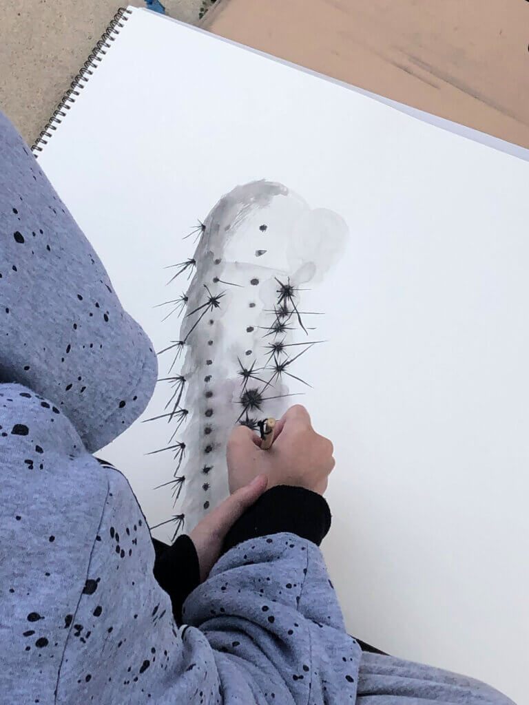 Photograph of a person painting a watercolor cactus with black spines in a spiral-bound sketchbook.