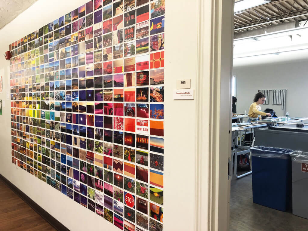 Photograph of a hallway installation made from hundreds of small color images mounted in a large grid beside an open classroom door.