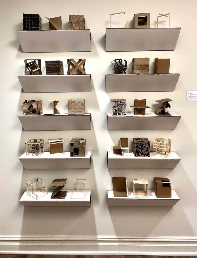 Wall display of small cardboard and wood sculptural models arranged in rows on floating white shelves.